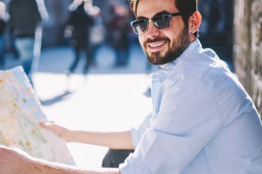 Şık güneş gözlüklü başarılı turistin portresi elinde seyahat haritası tutarken kameraya gülümsüyor ve şehir merkezindeki caddede oturan mimari gösterilerin doğru hedefini arıyor.