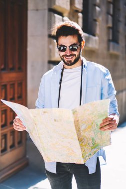 Pozitif sakallı turist, güneşli bir günde Kur 'an' da gezinerek haritada doğru yeri bulduğu için mutlu. Şık güneş gözlüklü neşeli erkek gezgin sokaklarda yürüyor.