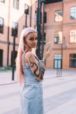 Büyük yönetim binasının iç avlusunda elinde cep telefonu olan dövmeli ve pembe saçlı bir kadının dikkatlice kameraya bakması.