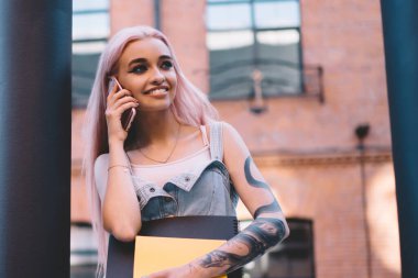 Sütunun yanında durup, şehir caddesinde hoş bir akıllı telefon konuşması yapan, gülümseyen ve yüzünü çeviren defterlerle mutlu genç bir kadın.