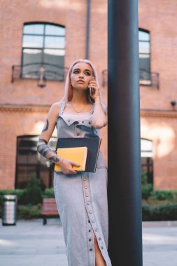 Pembe saçlı ve dövmeli ciddi genç bir öğrenci. Telefonlara cevap veriyor ve şehir caddesinde sütunun yanında dikilirken kameraya bakıyor.