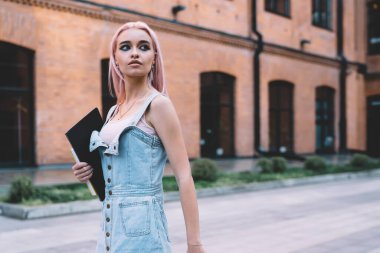 Pembe saçlı, elinde defterler ve akıllı telefonlar taşıyan ve kaldırımda yürürken yüzünü çeviren modaya uygun genç bir bayan.