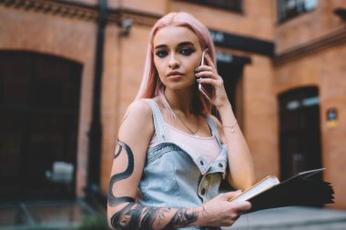 Yılan dövmeli, uzun pembe saçlı, kot elbiseli, cep telefonu rehberinden konuşan zarif bir bayan.