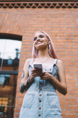 Dövmeli, uzun pembe saçlı, kot elbiseli, caddede cep telefonunda sörf yapan modern, neşeli bir bayan.