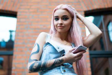 Düşük açılı, güler yüzlü, yılan dövmeli, uzun pembe saçlı, kot elbiseli, sokakta cep telefonuyla mesajlaşan bir kadın.