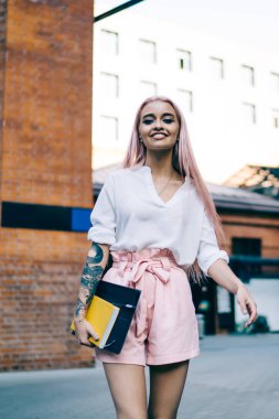 Beyaz gömlekli, pembe şortlu, uzun pembe saçlı, sıska gülümseyen kadın elinde kitap ve klasörle sokakta yürüyor.