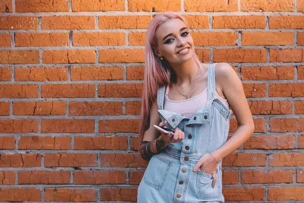 Akıllı telefonlu, kendine güveni tam genç bir kadın. El ele tutuşup kameraya gülümserken şehir sokaklarında tuğla duvara yaslanmış duruyor.