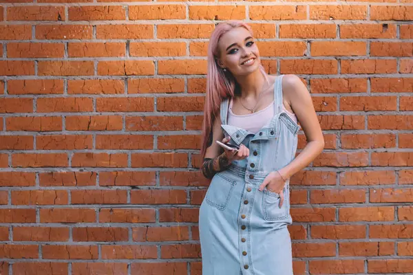 Akıllı telefonlu, kendine güveni tam genç bir kadın. El ele tutuşup kameraya gülümserken şehir sokaklarında tuğla duvara yaslanmış duruyor.