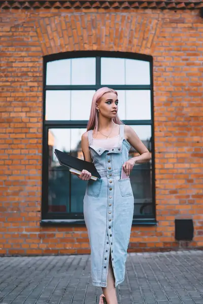 Elinde not defterleri olan modaya uyan genç bir kadın şehir caddesinde tuğla binanın önünde dururken akıllı telefonu cebine koyuyor.