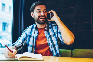 Pozitif sakallı hippi adam smartphone aygıt üzerindeki telefon görüşmesi sırasında gülüyor. Uzay coworking oturan telefonda konuşurken Not Defteri'nde bilgi yazma rahat kıyafet içinde neşeli erkek blogger
