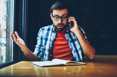 Ders kitabı tahta masada oturan dijital smartphone aygıt arama gözlük şok sakallı genç adam. Şaşırmış gibi yenilikçi öğrenci modern telefonla konuşurken rahat kıyafet giymiş