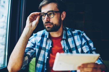 Uzağa bakıyor ve kendi blog geliştirmek için fikirleri üzerinde düşünme dokunmatik yüzey ile düşünceli sakallı genç adam. Gözlük vizyon düzeltme modern tablet elinde tutan durulması hipster adam