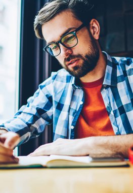 Dalgın sakallı öğrenci ödev Not defteri alanı coworking eğitim aşağı yazma. Akıllı hipster adam ders notları not defterinde kafede sınava hazırlık sırasında yapma gözlük