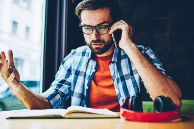 Kızgın genç adam kitap alan coworking içinde vasıl okul sırası oturan smartphone aygıt üzerinde iletişim gözlük sakallı. Telefon görüşmesi sırasında konuşurken rahat kıyafet rahatsız hipster adam giymiş