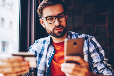 Aramak için gözlük kartvizit elinde tutan ve akıllı telefon numarası çevirmeden vizyon düzeltme için genç adam sakallı. Dalgın yakışıklı erkek blogger telefon hakkında bilgi arama