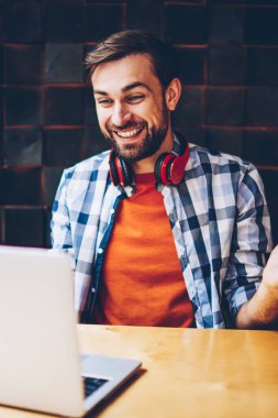 Mutlu sakallı hippi blogger modern dizüstü bilgisayarda görüntülü görüşme yoluyla arkadaşlarınızla iletişim kurmak için kablosuz 4 g bağlı internet. Komik eğitsel bir netbook izlerken boyun kulaklık ile pozitif genç adam