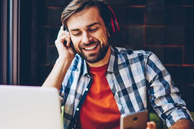Mutlu sakallı genç modern kulaklık smartphone aygıtında indirilen çalma listesinden müzik dinleme adam. Dijital dizüstü bilgisayar başında oturan radyo web sitesinde ses şarkı zevk neşeli erkek blogger