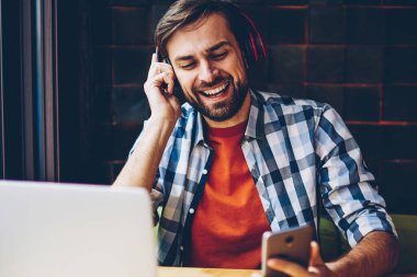 Pozitif sakallı genç adam favori ses çalma listesinden şarkıyı modern Kulaklığımda Smartphone'da indirilebilir. Mutlu hipster adam dinleme müzik radyo oyuncu laptop oturan telefonu