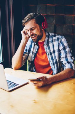 Smartphone için Bluetooth kaygısız sakallı genç adam şarkı şarkı dinleme ses modern kulaklık girişi sırasında bağlı. Neşeli erkek blogger dijital dizüstü bilgisayar kafede oturan müzik keyfi
