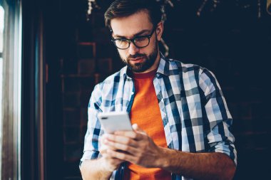 Dalgın sakallı hippi blogger gelen bildirim okuma ve ağlar Smartphone cep telefonu ile sohbet bağlı 4 g için Internet. Genç adam cep telefonu online video izlerken rahat gömlek giymiş