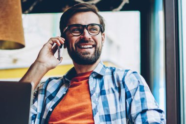 Smartphone üzerinde arama ve pencereden dışarı bakıyor telefon görüşme sırasında gülüyor neşeli sakallı hippi adam. Netbook çalışırken arkadaşıyla telefonda konuşurken olumlu erkek blogger