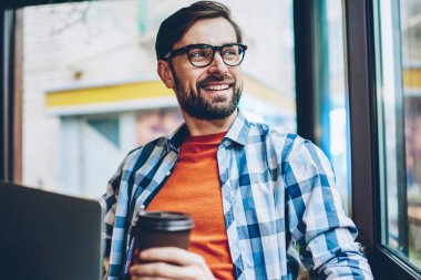 Gülümseyen hayal pencereden dışarı bakıyor ve öğle arasında Web sitesi geliştirmek için yaratıcı fikirler üzerinde düşünme genç adam sakallı. Düşünceli neşeli erkek blogger ile netbook aygıt çalışma gitmek için kahve