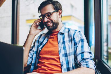 Happy sakallı genç adam gözlük smartphone yazılım yolu ile radyo 4 g modern dizüstü bilgisayarda güncelleştirilirken iletişim internet. Netbook hücresel oturma konuşurken olumlu hipster adam