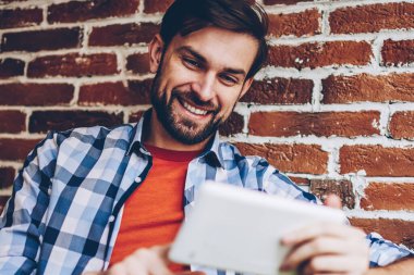 Mutlu sakallı genç adam modern Dokunmatik ped cihaz üzerinde Web sitesinde komik Haberler okuma rahat gömlek giymiş. Neşeli hipster blogger yayın 4 g internet bağlantısı üzerinden dijital tablet İndiriyor