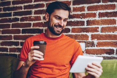 Gülümseyen sakallı erkek bogger kahve paket servisi olan restoran izlerken komik canlı stream sosyal ağ üzerinden Internet dijital tablet, e-posta dokunmatik yüzey üzerinde iyi haber okumak pozitif genç adam bağlı 4 g ile