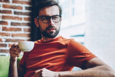Dalmış genç adam lezzetli kahve elinde tutan ve pencereden dışarı bakıyor 20 yaşında sakallı. Düşünceli hipster adam gözlük vizyon düzeltme kafede oturan içecek zevk için