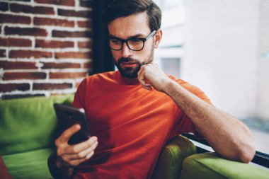 Sosyal ağlar üzerinde modern smartphone aygıt 4 g internet bağlantısı üzerinden video izlerken sakallı hippi blogger yoğunlaşmıştır. Dalgın genç adam üstünde hareket eden telefon aygıt Web sitesinde haber okuma