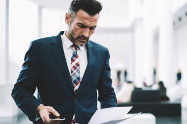Dalgın ciddi girişimci dikkatle finansal raporlar ofiste ayakta okuma takım elbise giymiş. Olgun işadamı smartphone ile kağıt belgeleri kontrol elinde konsantre