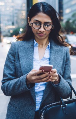Kafası meşgul, siyah çantalı, gri ceketli, yuvarlak gözlüklü, esmer, genç bir kadın New York 'ta yaya geçidinin yanındaki akıllı telefona dokunuyor.