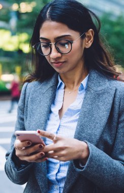 Gözlüklü, siyah çantalı ve gri ceketli, New York 'un kalabalık caddesinde, bulanık arka planda yaya geçidinde akıllı telefonlara tıklayan ciddi şekilde şık, genç, esmer bir kadın.