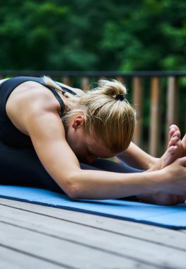 Genç, zayıf, sarışın kadın, esneme egzersizleri yapıyor. Mavi paspasın üzerinde el ele tutuşarak bacaklara korpus uzatıyorlar.