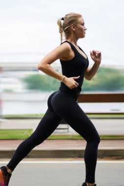 Siyah eşofmanlı güzel zayıf vücutlu, kendinden emin bir sporcunun yan görüntüsü yaz parkındaki bulanık sahil şeridine doğru ilerliyor.