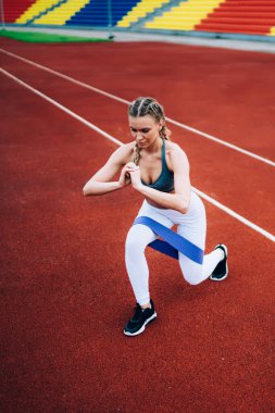 Spor kıyafetleri ve spor ayakkabıları giyen konsantre sarışın kadın stadyumdaki antrenman sırasında lastik kurdeleyle egzersiz yapıyor ve göğsüne yakın elle saldırıyor.