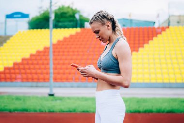 At kuyruklu ve ciddi yüzlü, yürüyüş pistinde duran ve sosyal medyada mesaj okuyan güzel, aktif bir kadının yan görüntüsü