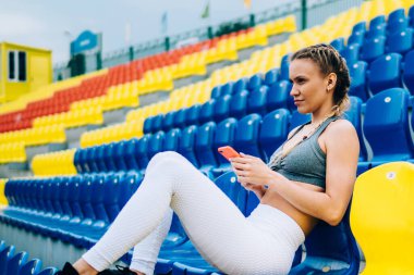 Beyaz tozlu sarı saçlı ve gri bluzlu kararlı bayan sporcu stadyumda akıllı telefonlardan sörf yaparken oturuyor ve gözlerini kaçırıyor.