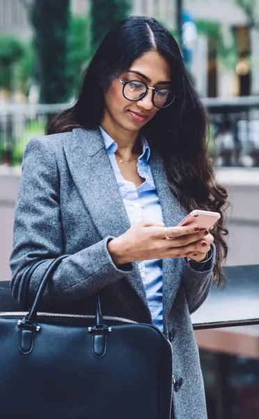 Gözlüklü, gri ceketli, deri çantalı ve cep telefonuyla mesajlaşan havalı etnik kadın şehrin arka planında duruyor.