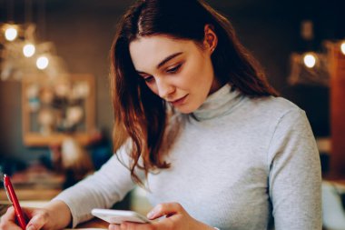 Ciddi kadın serbest çalışan not defterinde cep telefonuyla iletişim kuran, birlikte çalışma alanında çalışan, teknolojiyi ve bilgi aramak için kablosuz bağlantıyı kullanan genç bir kadın.