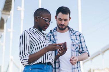 Kısa saçlı, gözlüklü siyahi bir kadın akıllı telefon ekranı gösteriyor. Köprüde günlük kıyafetlerle yürüyen ciddi bir genç adam.