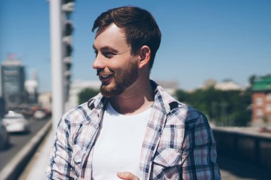 Keyifli, modern, sakallı, normal tişörtlü ve kot pantolonlu bir erkek yaz günü köprüde yürürken akıllı telefon uygulaması kullanıyor.