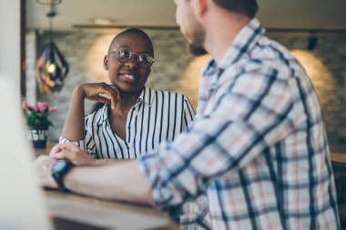 Yandan bakan genç sakallı adam, kafeteryada dizüstü bilgisayarlı cam içinde oturan neşeli Afrikalı Amerikalı kadınla konuşuyor.
