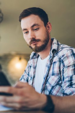 Kafede dizayn edilmiş iç dizaynı olan bir masada dizüstü bilgisayarla çalışırken gömlekli, sakallı ciddi bir adamın cep telefonuyla eğlenmesi.