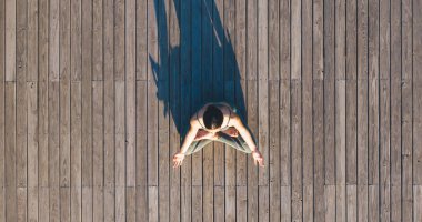 Spor kıyafetli kadın, Lotus pozisyonunda otururken ahşap zeminde yoga yapıyor ve gölgesi arkadan düşüyor.