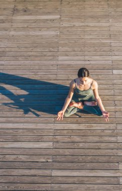 Yoga kıyafetli genç bir kadın ahşap zeminde lotus pozisyonunda oturuyor ve gölgesi düşerek meditasyon yapıyor.