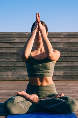 Odaklanmış, spor kıyafetli, mindere oturmuş nilüfer çiçeği ve Namaskar pozu vererek yoga yapan, ahşap gezinti güvertesinde sakin ve sağlıklı bir yaşam süren zayıf bir kadın.