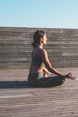 Nilüfer pozisyonunda meditasyon yapan, elleri dizlerinde, gözleri tahta kaldırımda kapalı, dingin, dişi yogi 'nin yan görüntüsü.