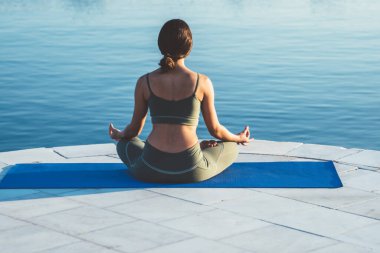 Tanımlanamayan sakin bir kadının deniz manzarası önünde mavi mindere oturup dinlenirken yoga yapması ve meditasyon yapması.